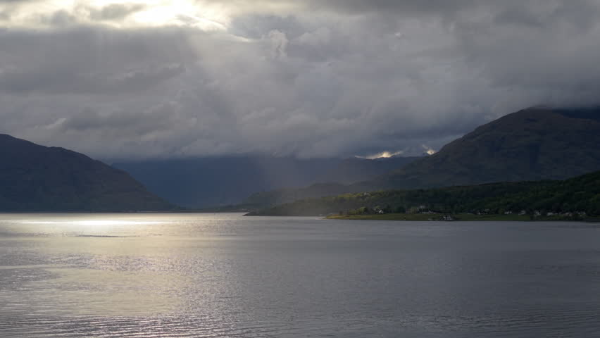 Dramatic drone view over Loch Linnhe in the afternoon, showing sunbeams bursting through heavy storm clouds to illuminate the Scottish Highland shoreline