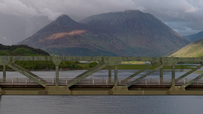 Telephoto drone tracking shot compressing the green steel truss of the Ballachulish Bridge against the dramatic, cloud-capped mountains of Glencoe.