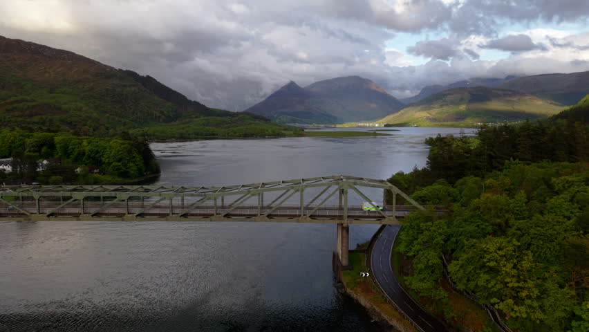Shallow drone orbit around the Ballachulish Bridge, showcasing its steel truss structure against the expansive, sun-dappled mountains of Glencoe.