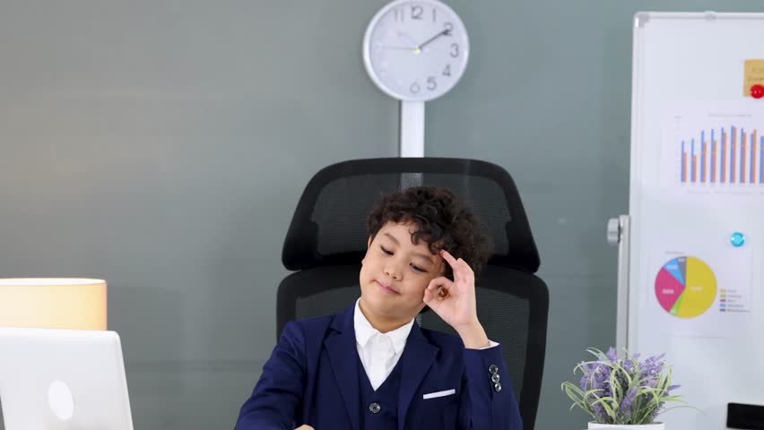 Young boy in a blue suit makes an okay sign while sitting at a desk