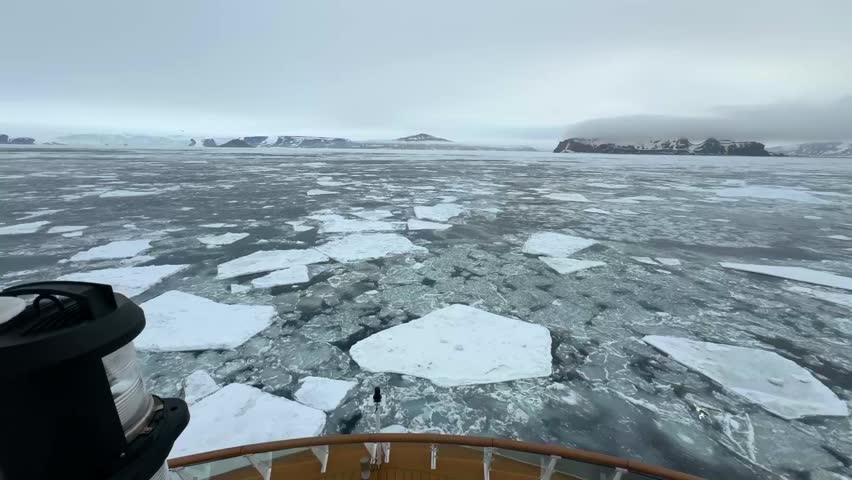 Expedition ship moves through fragmented pack ice in the Weddell Sea surrounded by frozen Antarctic waters and distant snow covered islands, static forward facing polar navigation shot.