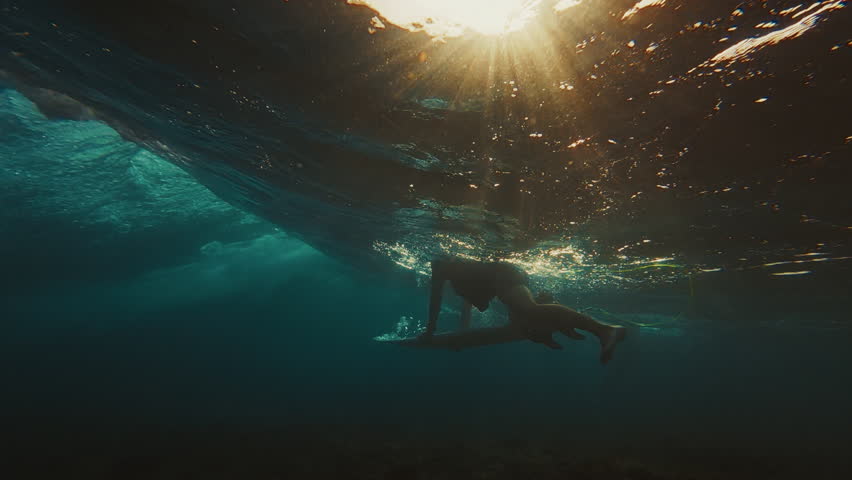 Surfer dives under the ocean wave with surfing board and passes it. Underwater view of the surfer doing duck dive trick