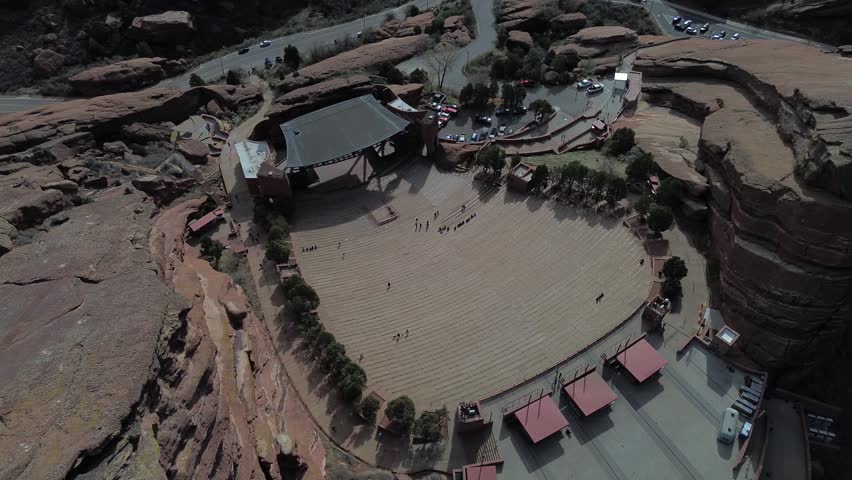 Stage canyon perspective highlights Red Rocks seating bowl near Alameda Parkway with massive sandstone fins rising around venue. Layered rock textures, scattered shrubs warm light, Morrison, CO. USA