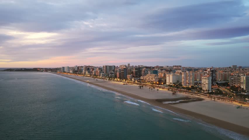 Illuminated city of Alicante and its long sandy beach during colorful blue hour dusk in Spain. Aerial drone rising, copy space
