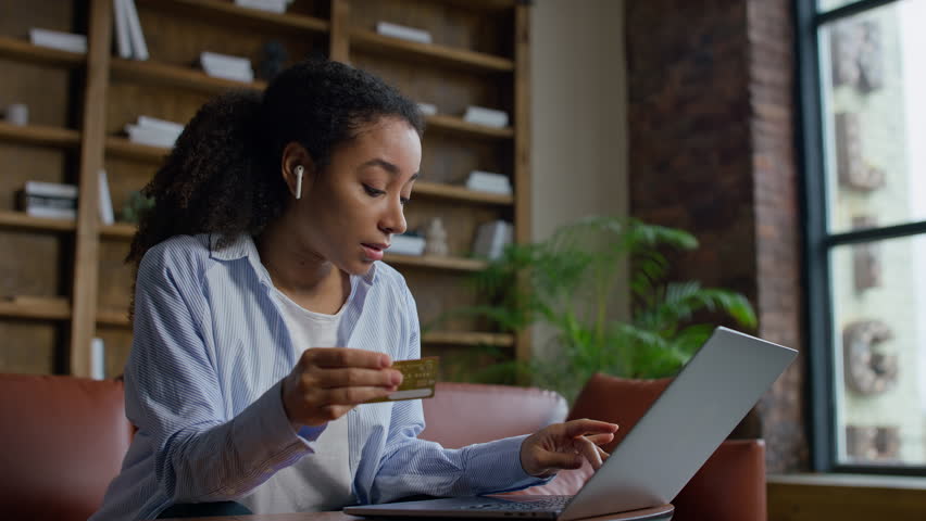 Young woman sitting on a sofa holding a credit card while using a laptop, showing online shopping, digital payment, and home banking