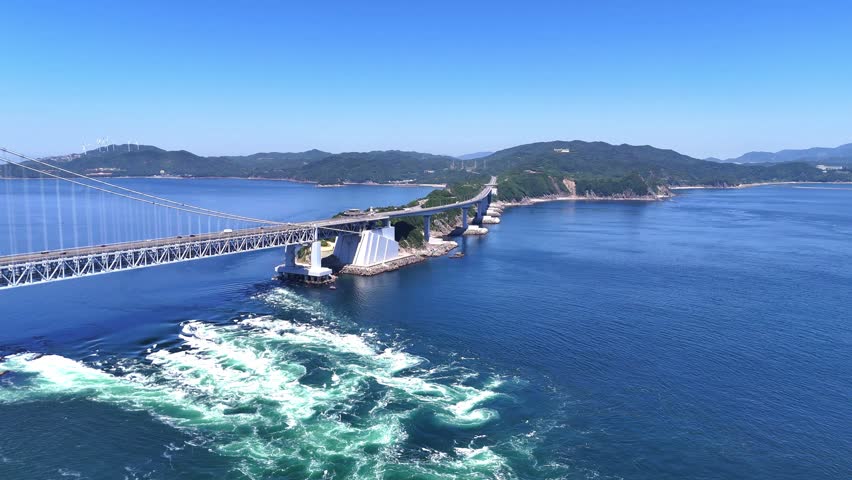 Spectacular aerial view of the Onaruto Bridge stretching across the beautiful blue ocean of the Naruto Strait, Japan. The iconic whirlpools can be seen swirling dynamically in the water below.

