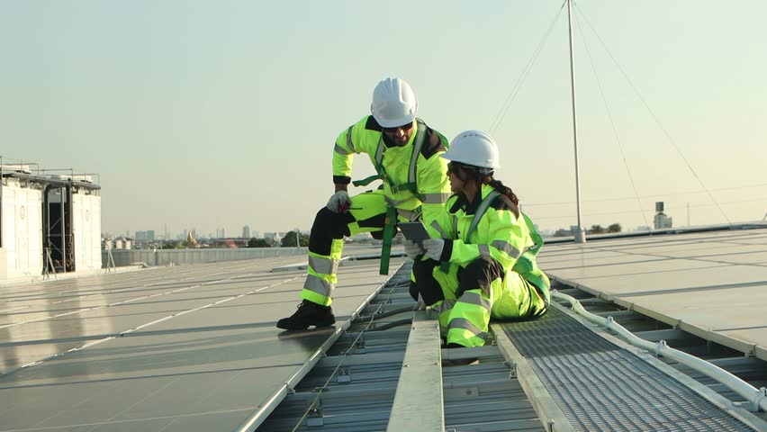 Two service engineers in safety clothing with helmet check solar system at rooftop solar farm with city view. Engineers installing solar panels. Man and woman workers inspecting solar panels on roof.