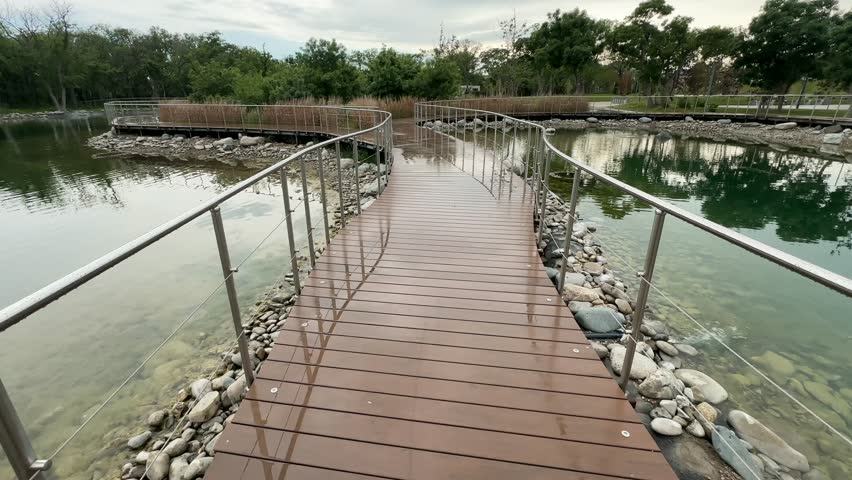 Park boardwalk forward motion over lake, curved wooden path, wet deck, railings, green trees, urban nature, travel recreation