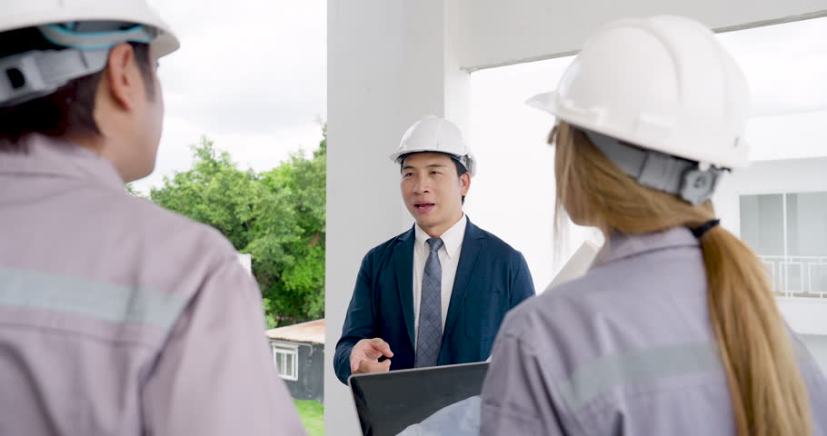 Asian male manager explains construction site safety inspection in slow motion while female engineer and contractor listen, supporting building project progress meeting inside unfinished site.