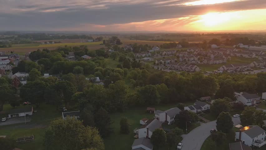 Aerial sunset view over a suburban neighborhood and rural farmland with golden evening light, rolling hills, trees, homes, and dramatic clouds in the countryside.