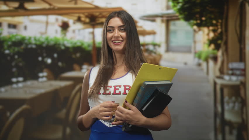 Young hispanic woman holding a yellow folder and books, hand to chest, smiling in a street cafe; optimism study.