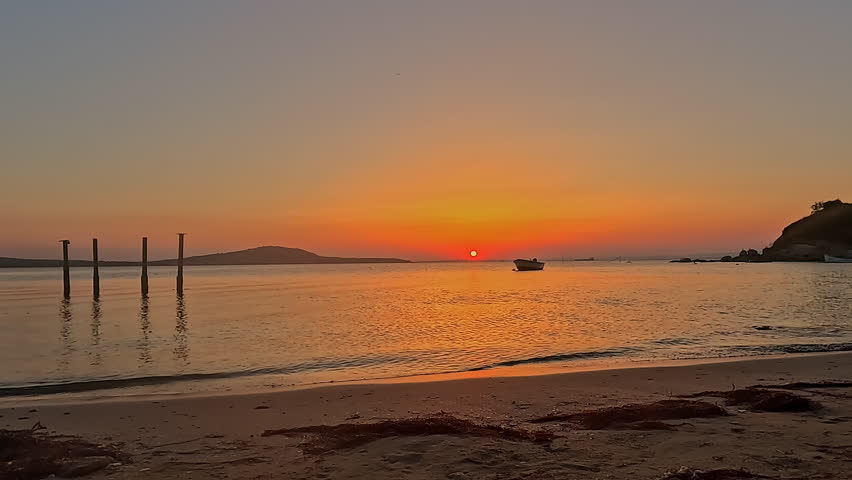 Peaceful summer sunset over calm sea with small fishing boat, sandy beach shoreline and warm orange sky. Scenic tropical vacation seascape background footage ideal for travel, relaxation and wellness themes.