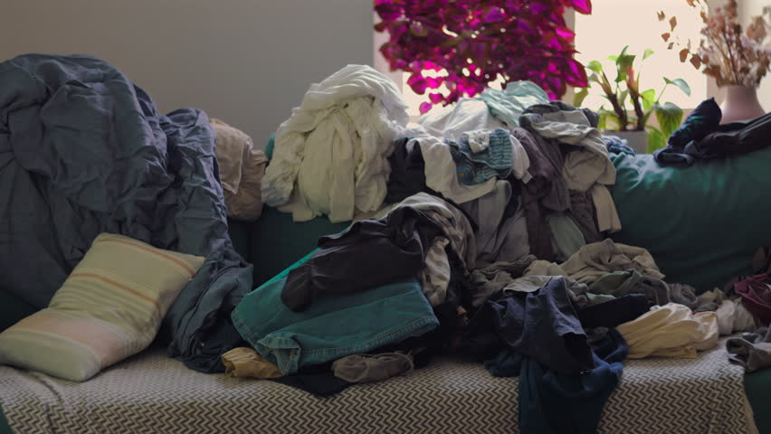 Panning view of messy couch filled with layered clothing and surrounding clutter highlighting buildup of laundry and everyday household pressure in quiet interior