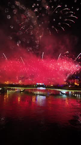 Massive red fireworks show illuminating the night sky and reflecting on the water surface, celebrating the vijit chao phraya festival over an urban cityscape with a bridge and a boat passing by, Bangkok, Thailand.