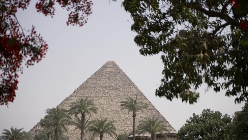 A cinematic view of the ancient Pyramid of Khafre in Giza, Egypt, beautifully framed by lush green foliage, blooming red flowers, and palm trees in the foreground.