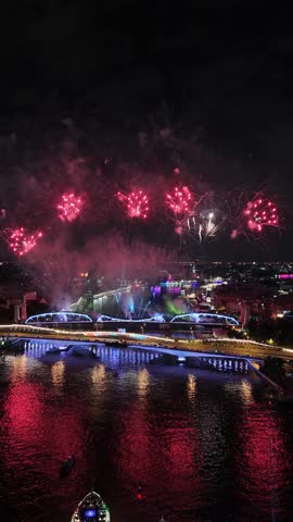 Colorful fireworks exploding in the night sky above an illuminated city bridge in Bangkok, Thailand, with tour boats watching the celebration from the river below, reflecting the vibrant lights on the water.