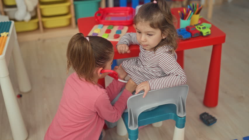 Two girls playing with a toy stethoscope at a red playtable, one holds hand to stomach in a building; gentle concern.