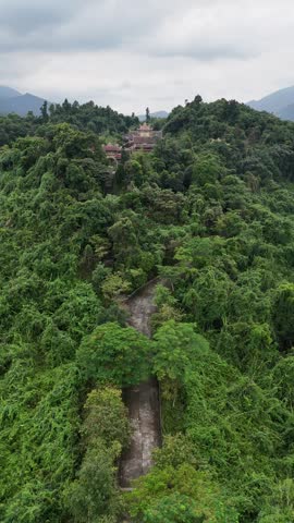 Drone view flying along a stone path leading to a buddhist temple hidden in a lush green jungle on a mountain in Hue, Vietnam, revealing a landscape with a lake and mountains in the background.