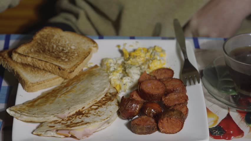 Person picking up a ham and cheese quesadilla from a hearty breakfast plate with fried sausage, scrambled eggs, and toast