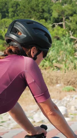 Portrait of young sportive female cyclist riding on road bicycle outdoors. Athletic woman in black helmet and cycling jersey cycling on bicycle lane along green forest. Vertical video