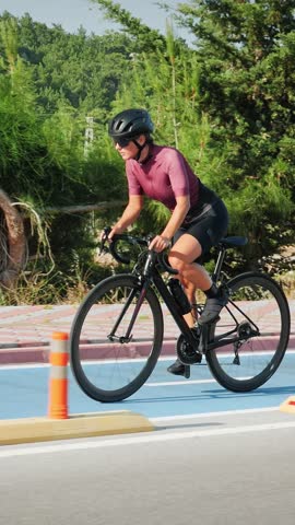 Female road cyclist riding on bike along asphalt bicycle lane road in sunshine. Young woman riding bicycle at sunset. Vertical video