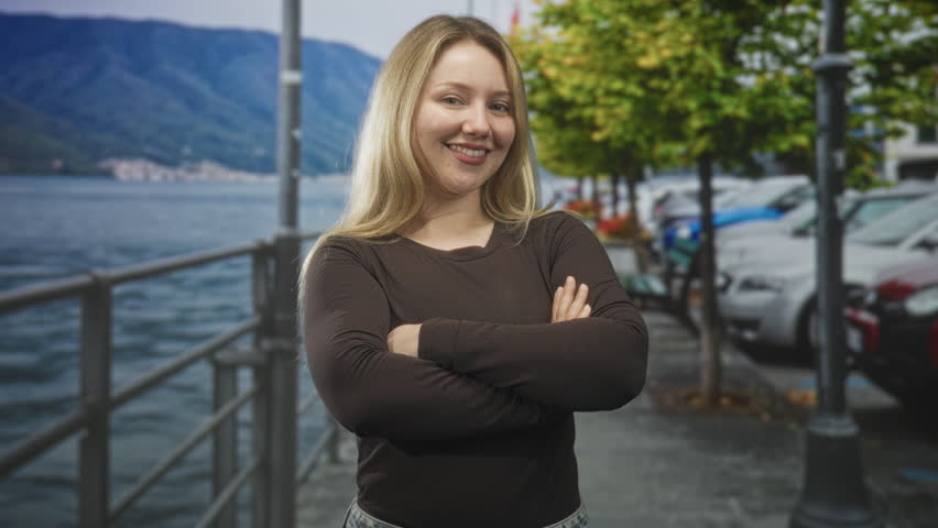 Woman with arms crossed smiling on a street waterfront promenade beside parked cars and metal railing; confidence approachability.