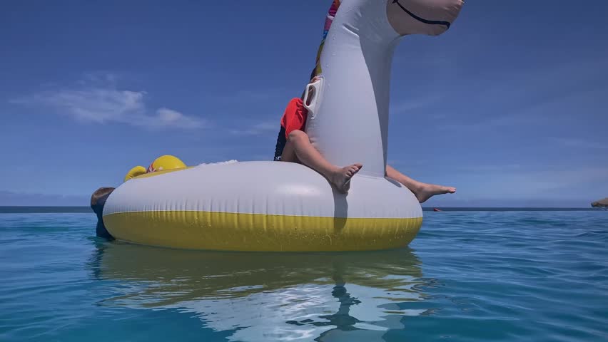 Child perched on inflatable unicorn, small silhouette against vast blue sky, toes dangling, lifevest visible, playful solo moment and confident balance on calm sea
