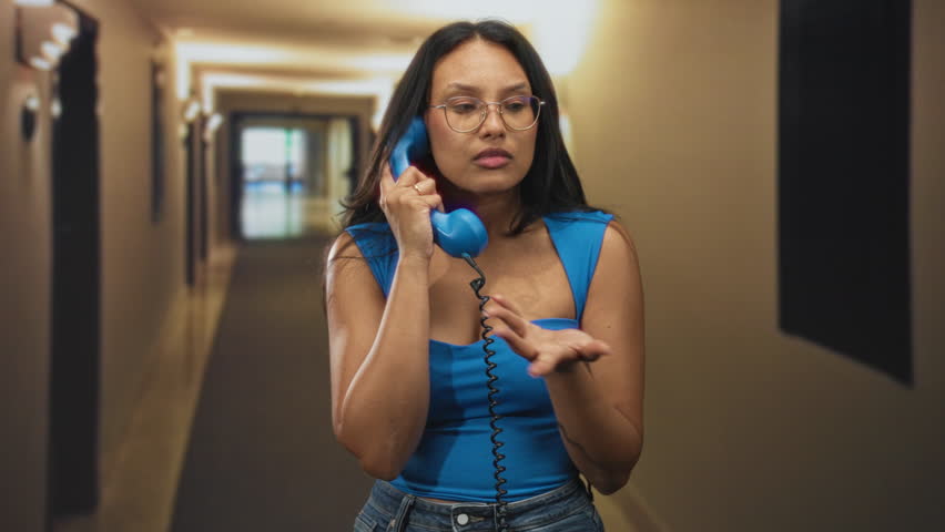 Young woman holding a vintage blue handset to her ear with hand in a hotel building corridor; quiet contemplation.