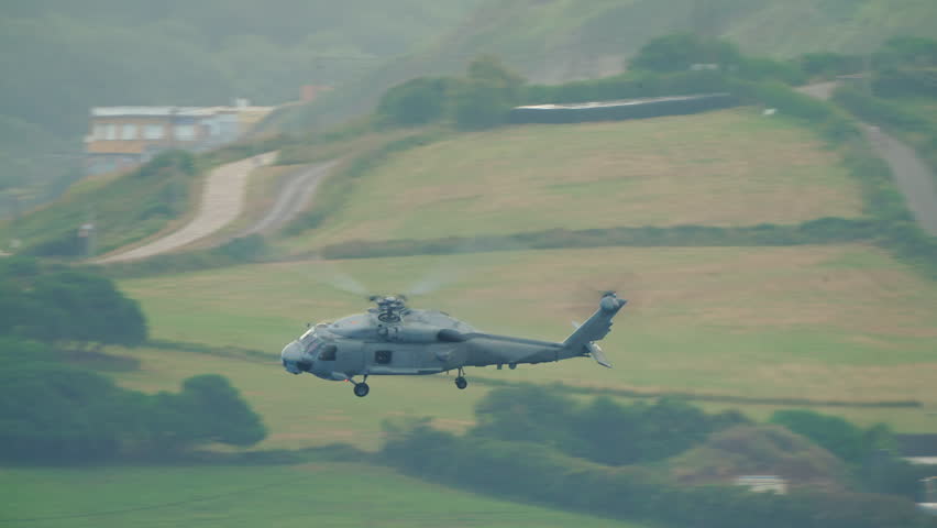 Large grey military helicopter passing at low altitude over a green coastal cliff with hazy sea and sandy beach visible in the distance