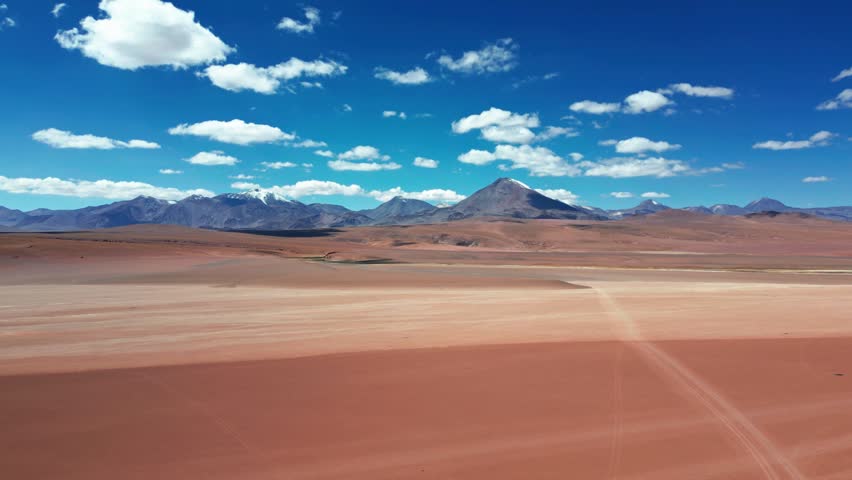 Slow backward flight over vast red arid altiplano desert plain with faint tire tracks revealing dramatic chain of volcanic peaks under vivid blue sky and clouds