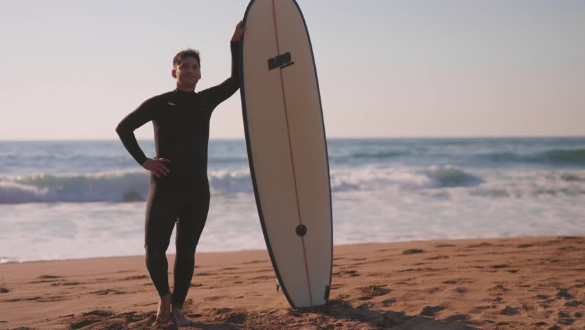 Portrait of a proud surfer wearing a wetsuit standing with his surfboard on the sandy beach during a beautiful sunset, getting ready for an evening surf session in the ocean waves