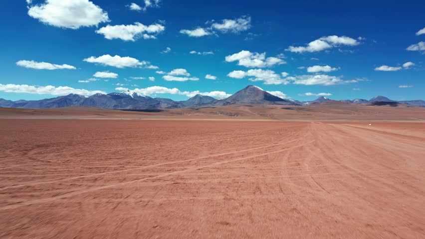 Fast low forward flight skimming over red arid altiplano desert floor with faint tire tracks leading directly toward dramatic chain of volcanic peaks under blue sky