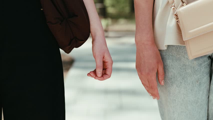 Two people hold hands while walking through a park. They are on a pathway with trees on either side, enjoying their time together on a sunny day