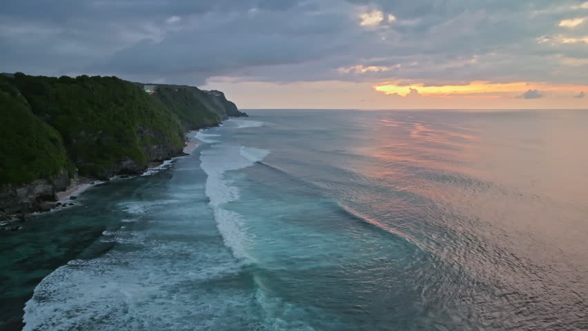 Bukit Peninsula coastline at Uluwatu with green cliffs meeting gentle ocean waves, glowing in warm golden hour sunset hues, tranquil scenic tropical seascape. Travel Bali. Aerial view drone flight