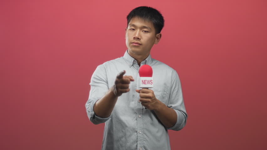 Man holds news microphone and points finger at camera in live studio with red backdrop; calm reporting.