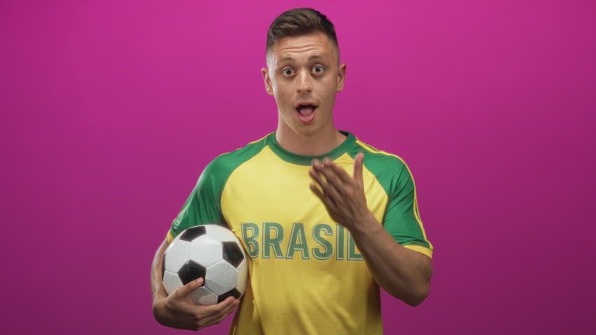 Man holding a soccer ball, hand covering mouth while smiling, wearing brasil jersey against magenta backdrop in studio; team pride playful.