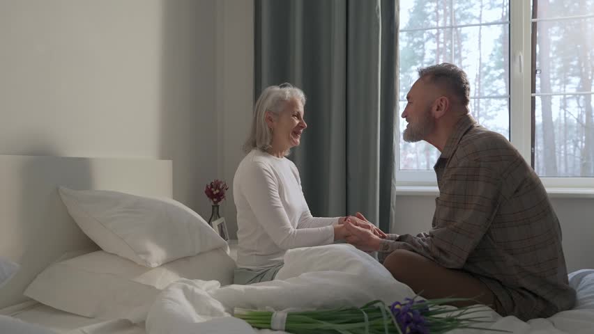 A man sits on a bed and talks while holding hands with a woman next to him