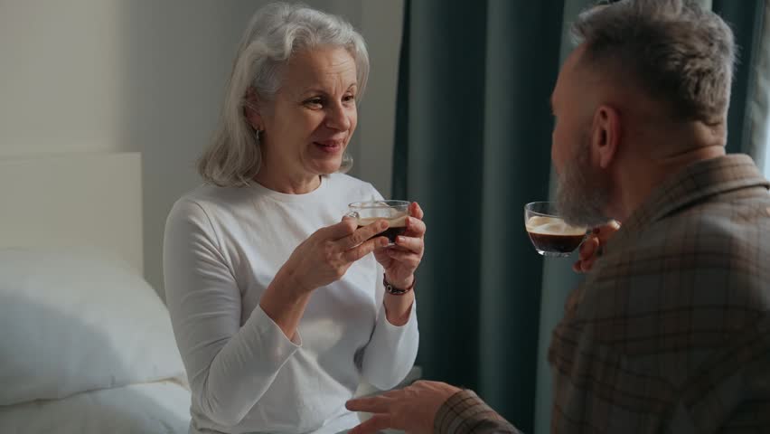 A woman and a man are holding cups while talking and smiling while sitting on the bed