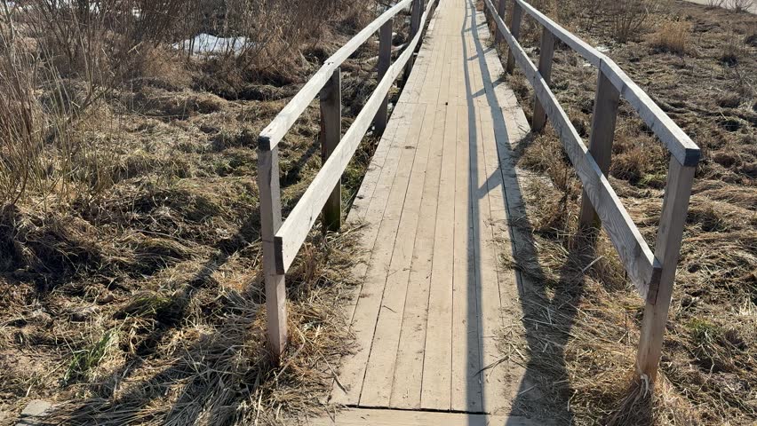 Temporary wooden bridge over dry grass, narrow plank walkway with handrails casting long shadows across dead brown vegetation