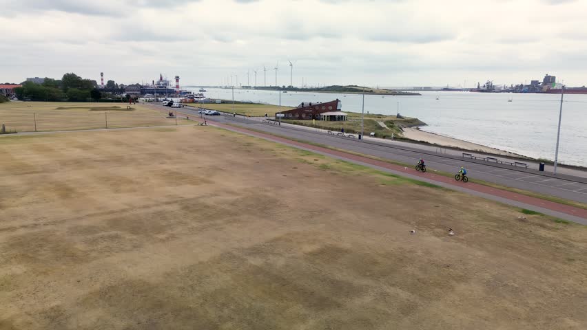 An aerial view shows a coastal road and bike path next to a wide grassy field. In the distance, port infrastructure, ships, and wind turbines stretch along the waterfront under a cloudy sky.