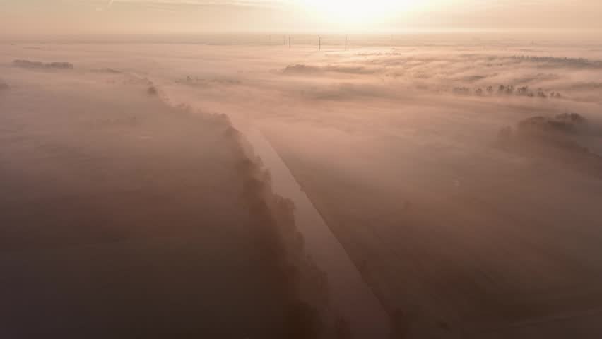 An aerial morning view shows thick fog rolling over fields and a calm river. Wind turbines break through the mist on the horizon under a soft, golden sunrise sky.