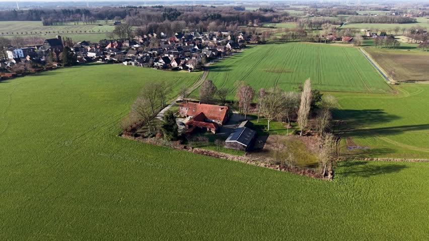 An aerial view shows a traditional brick farmhouse nestled among trees in the middle of vast green fields. A small residential village sits in the background under a clear sky.