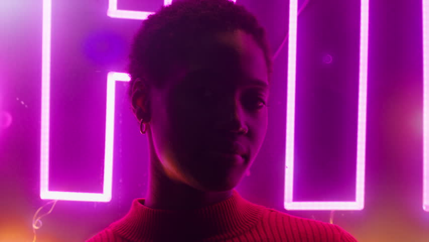 Young black woman in front of glowing neon lights at night. Colorful pink and purple illumination creates modern futuristic nightlife atmosphere