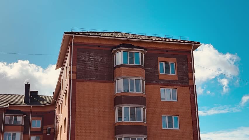 Sunlit brick apartment building under sky textured facade with curved balconies, multiple windows, weathered roofline and drifting