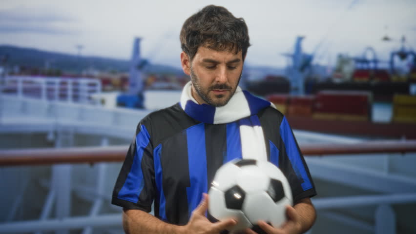 Man holding soccer ball, wearing blue striped team t shirt and scarf, and showing two fingers to face on a port ship deck; team pride.