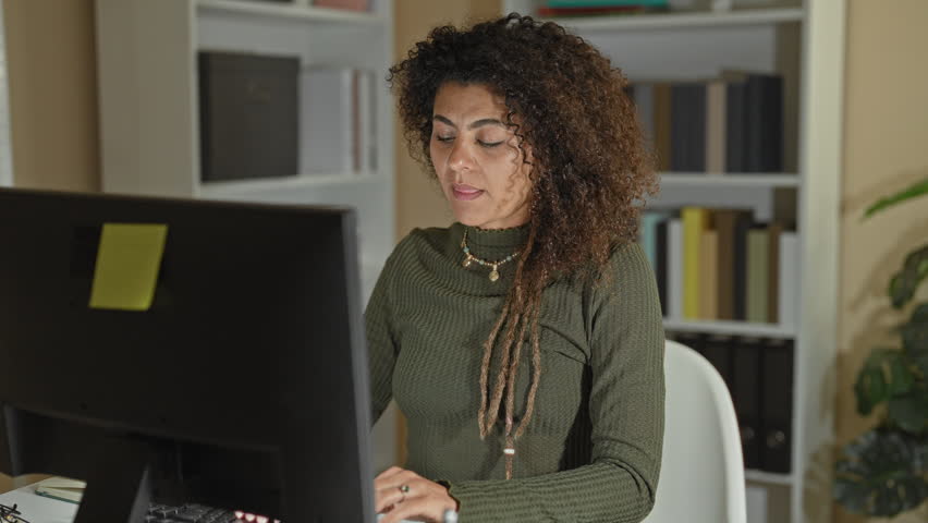 Woman rubs shoulder while using computer and mouse at desk in office building with monitor and keyboard visible; fatigue neck strain.