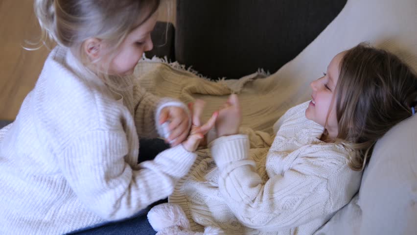 Two young sisters having fun together as one tickles the other while she lies on the couch, both laughing and enjoying a playful moment in the living room during a carefree day at home