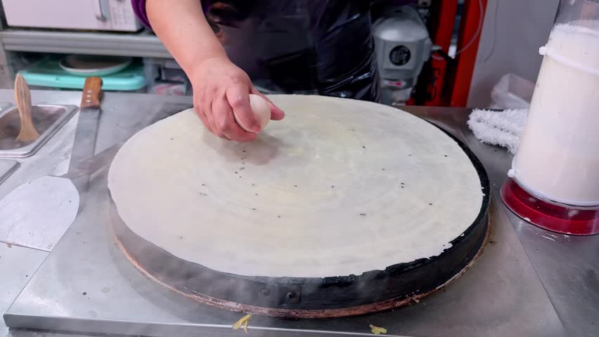 Close up of a street cook preparing a traditional Chinese jianbing, spreading a savory crepe on a hot griddle, adding egg and toppings in a bustling food stall in Chongqing, China