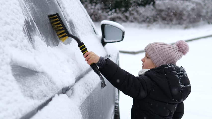 Cute little girl in a warm winter jacket and hat helping to brush fresh snow off a family car after a heavy snowfall, demonstrating responsibility and winter season chores during a cold day
