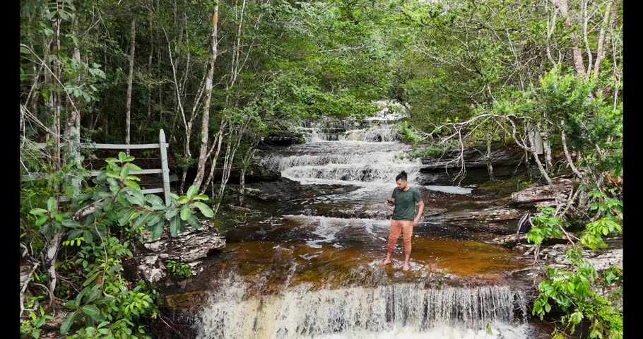 A person enjoys nature in the foreground, with a stunning waterfall cascading in a vibrant green setting in Colombia.